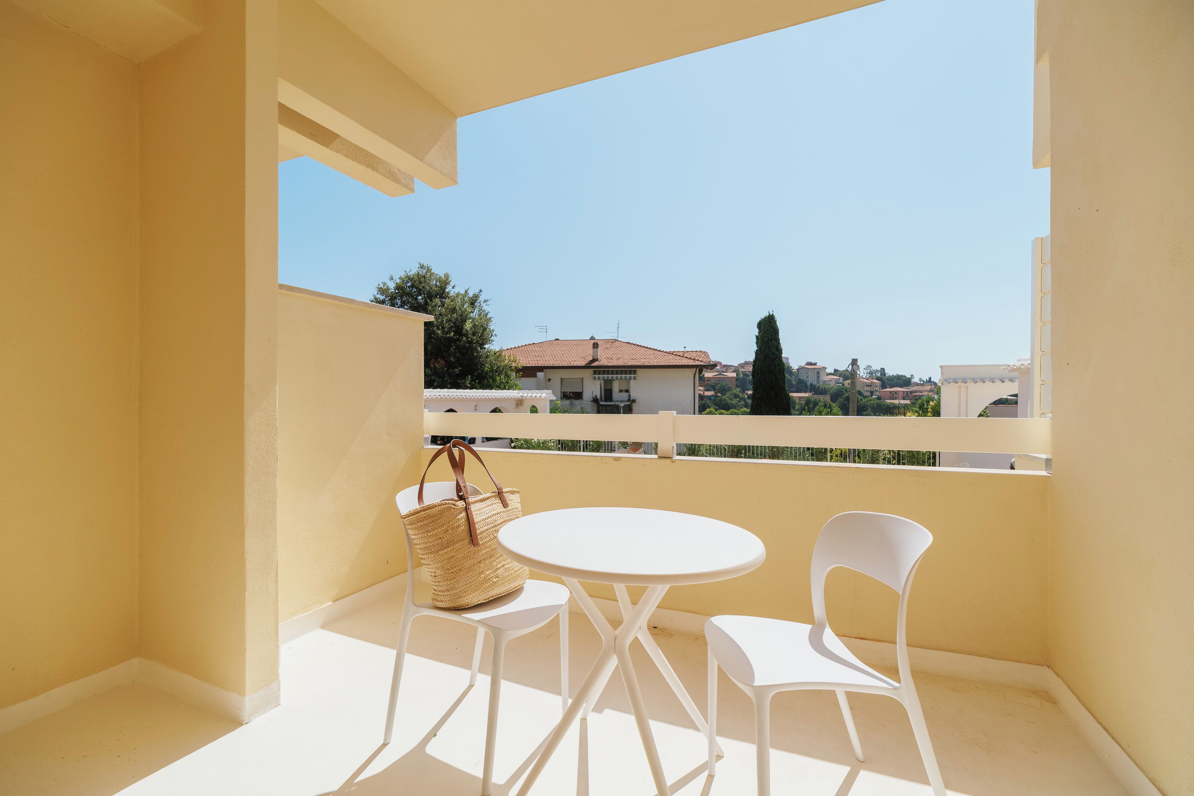 Un balcon avec une table blanche et deux chaises blanches, un panier en osier et une vue sur des bâtiments au loin.