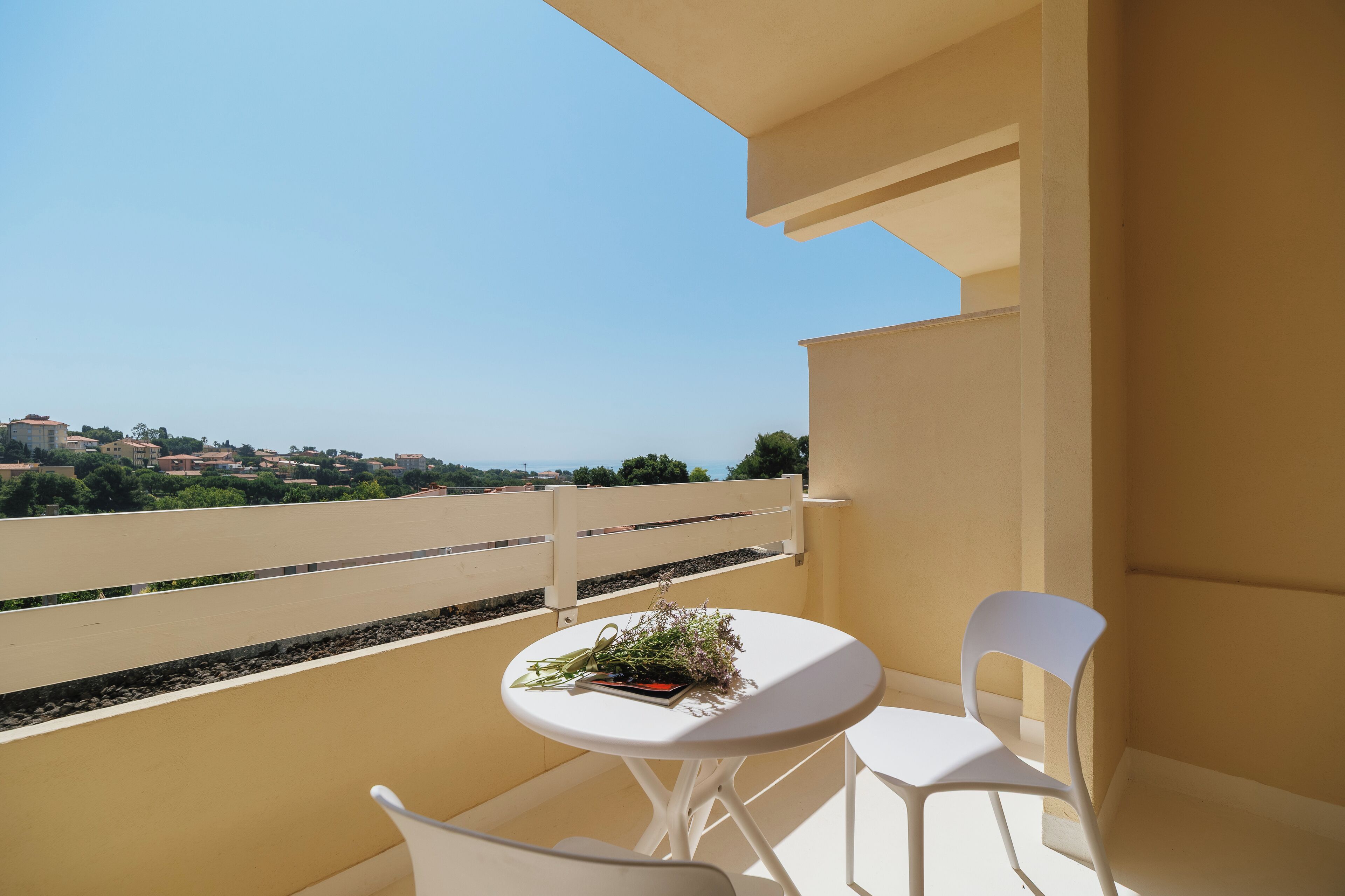 Un balcon avec une table blanche et deux chaises blanches, un petit bouquet de fleurs et une vue dégagée sur le paysage environnant.