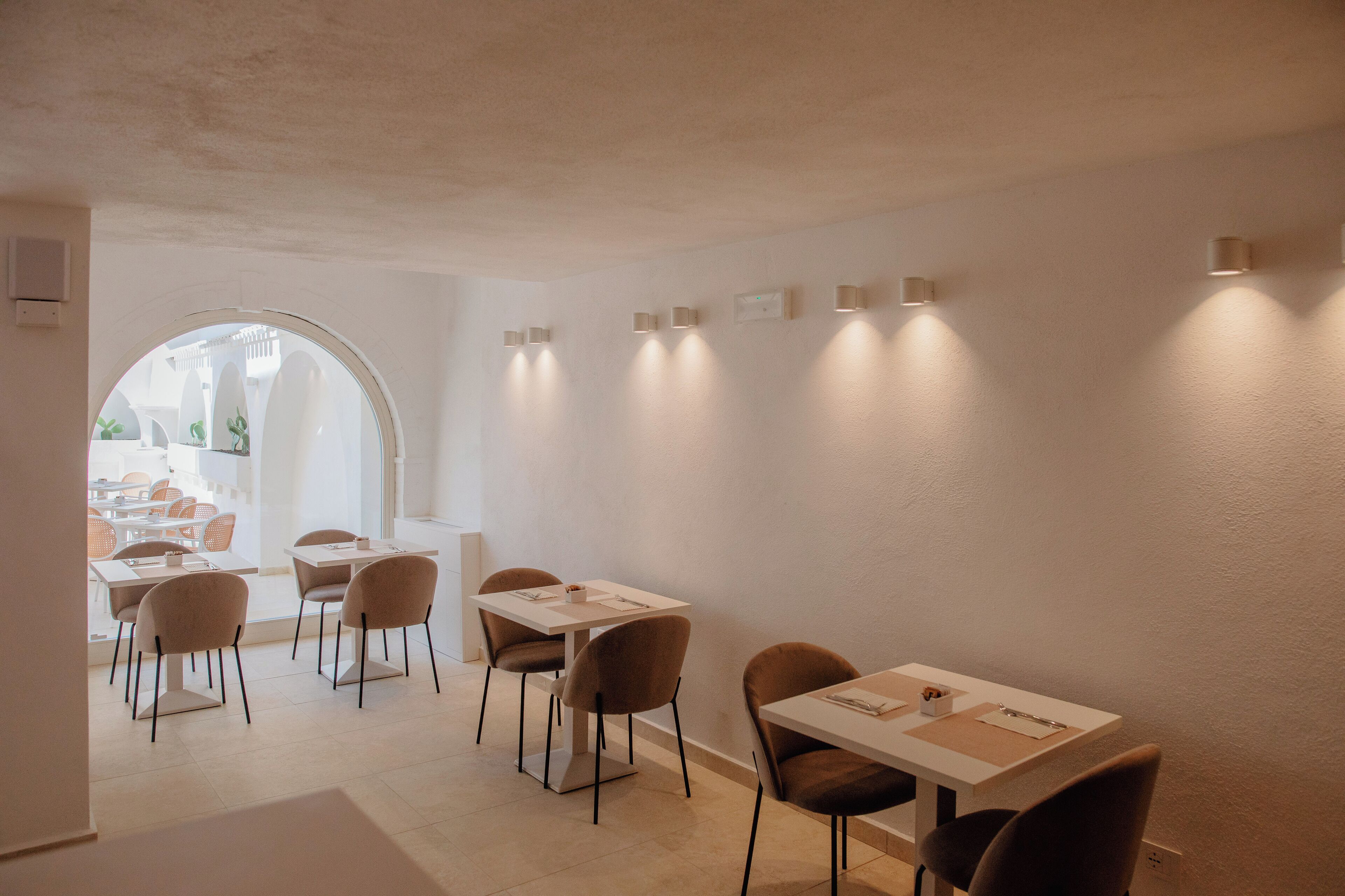 Comedor con mesas y sillas, un amplio ventanal arqueado e iluminación en la pared.