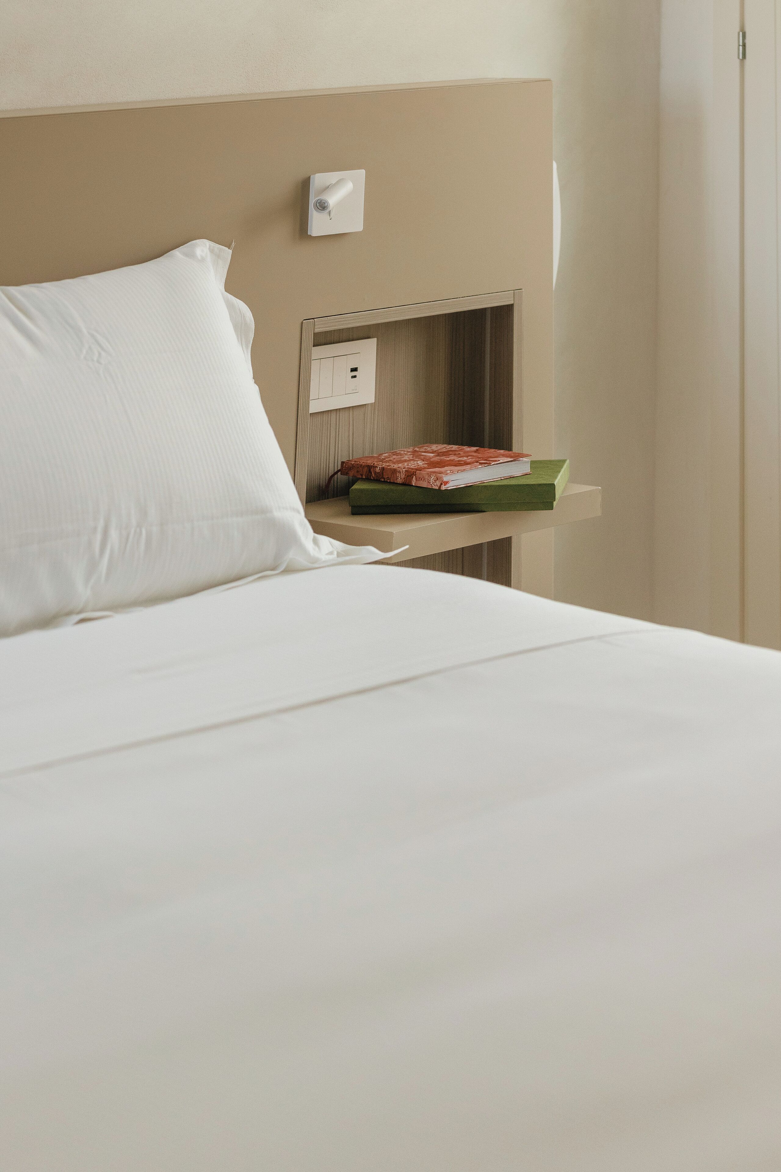 A neatly made bed with white linens, a pillow, and a small shelf with a book and a decorative item.