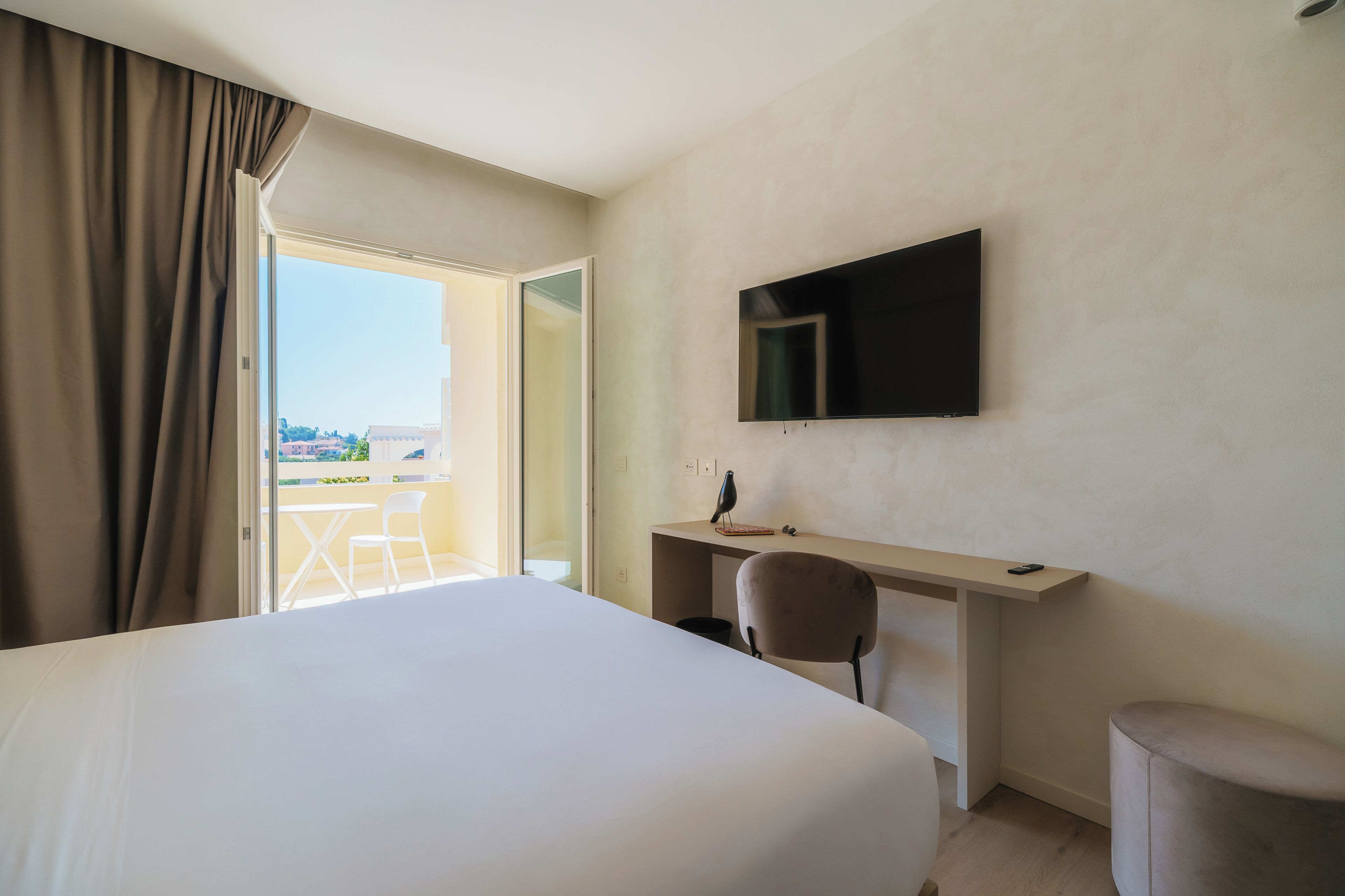 A hotel room with a large bed, a desk with a chair, a flat-screen TV mounted on the wall, and a view of the outdoors through an open door.