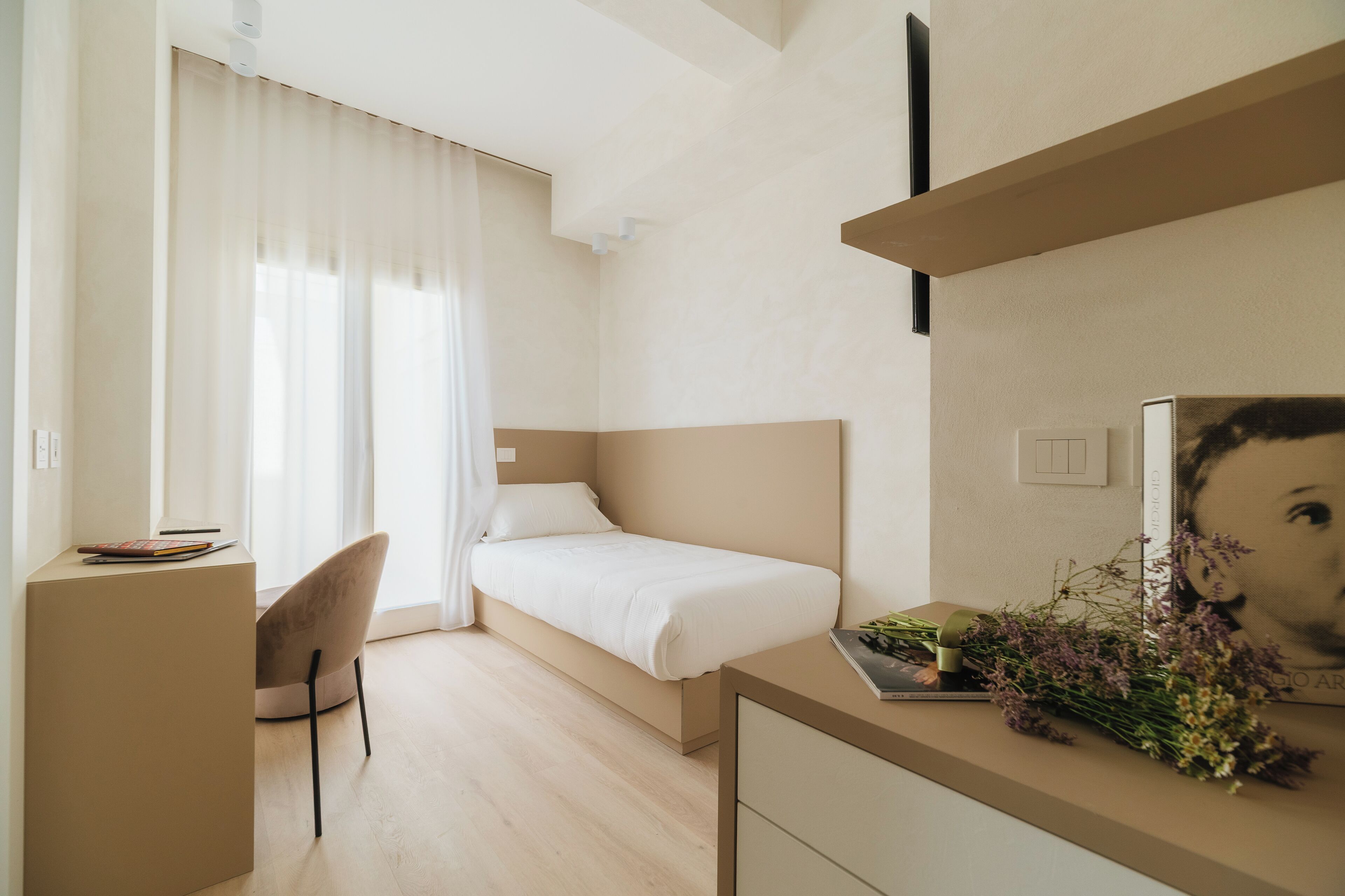 A modern hotel room with a bed, a desk, a chair, and a small table with flowers.