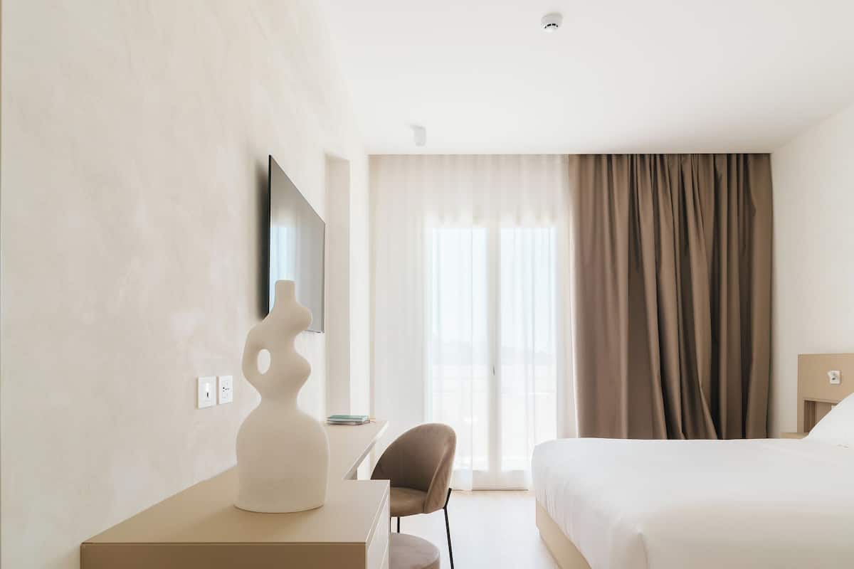 A modern hotel room with a large bed, a flat-screen TV mounted on the wall, a small seating area, and a large window with curtains.