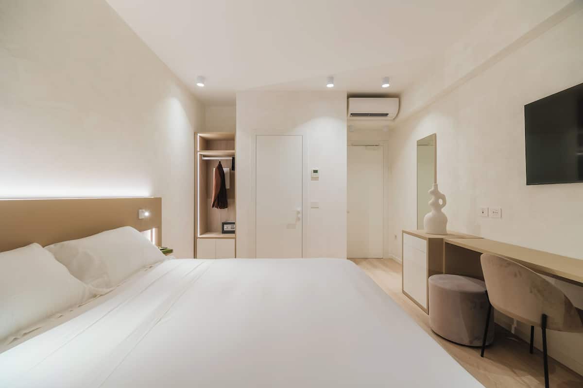 A modern hotel room with a large bed, a desk with a chair, a wardrobe, and a mirror.