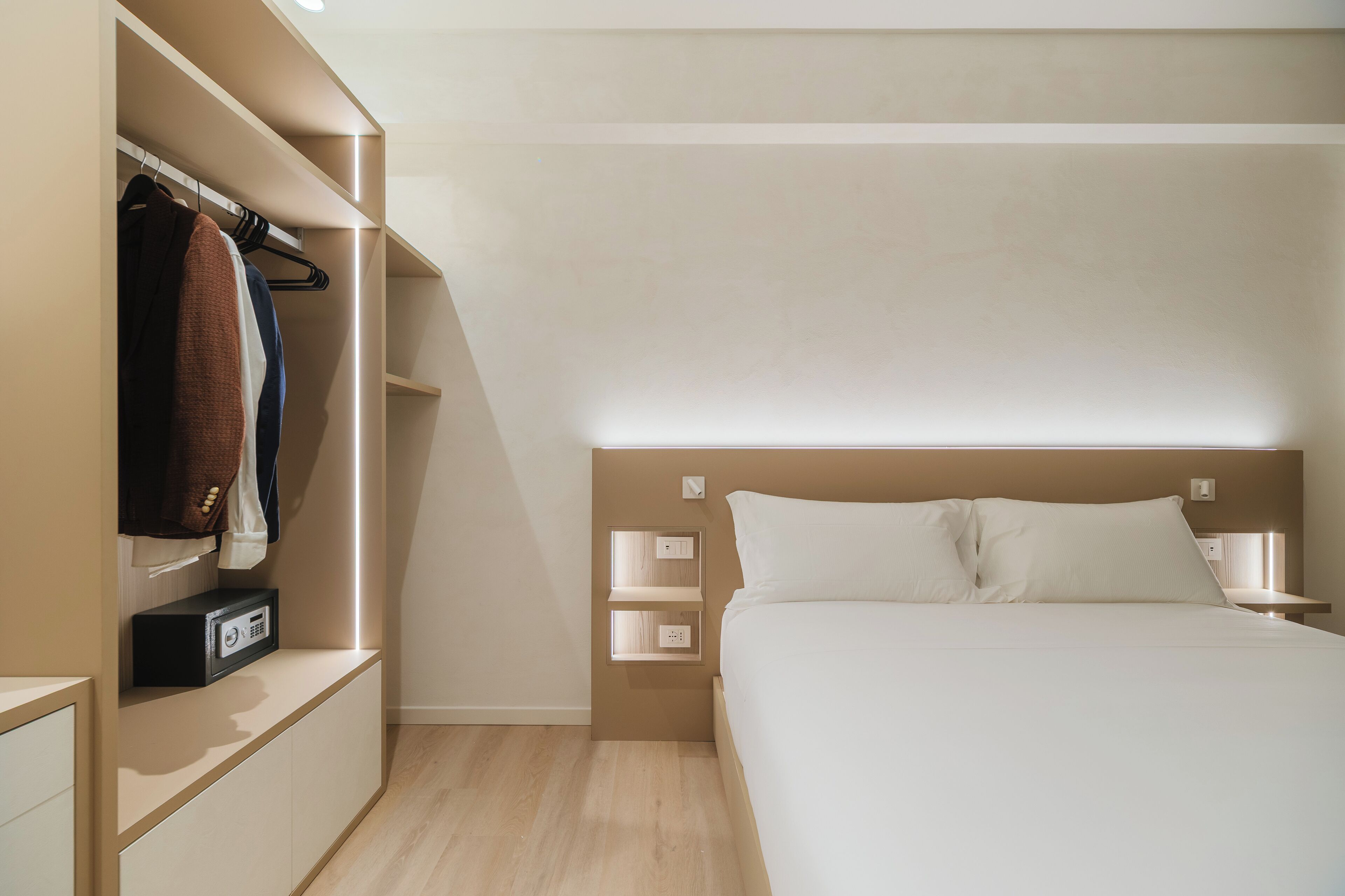 A modern hotel room with a bed, bedside tables, a wardrobe, and built-in lighting.
