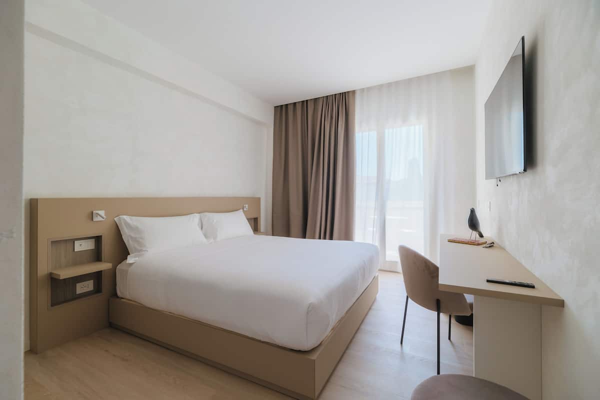 A hotel room with a bed, bedside tables, a desk, a chair, and a TV mounted on the wall.