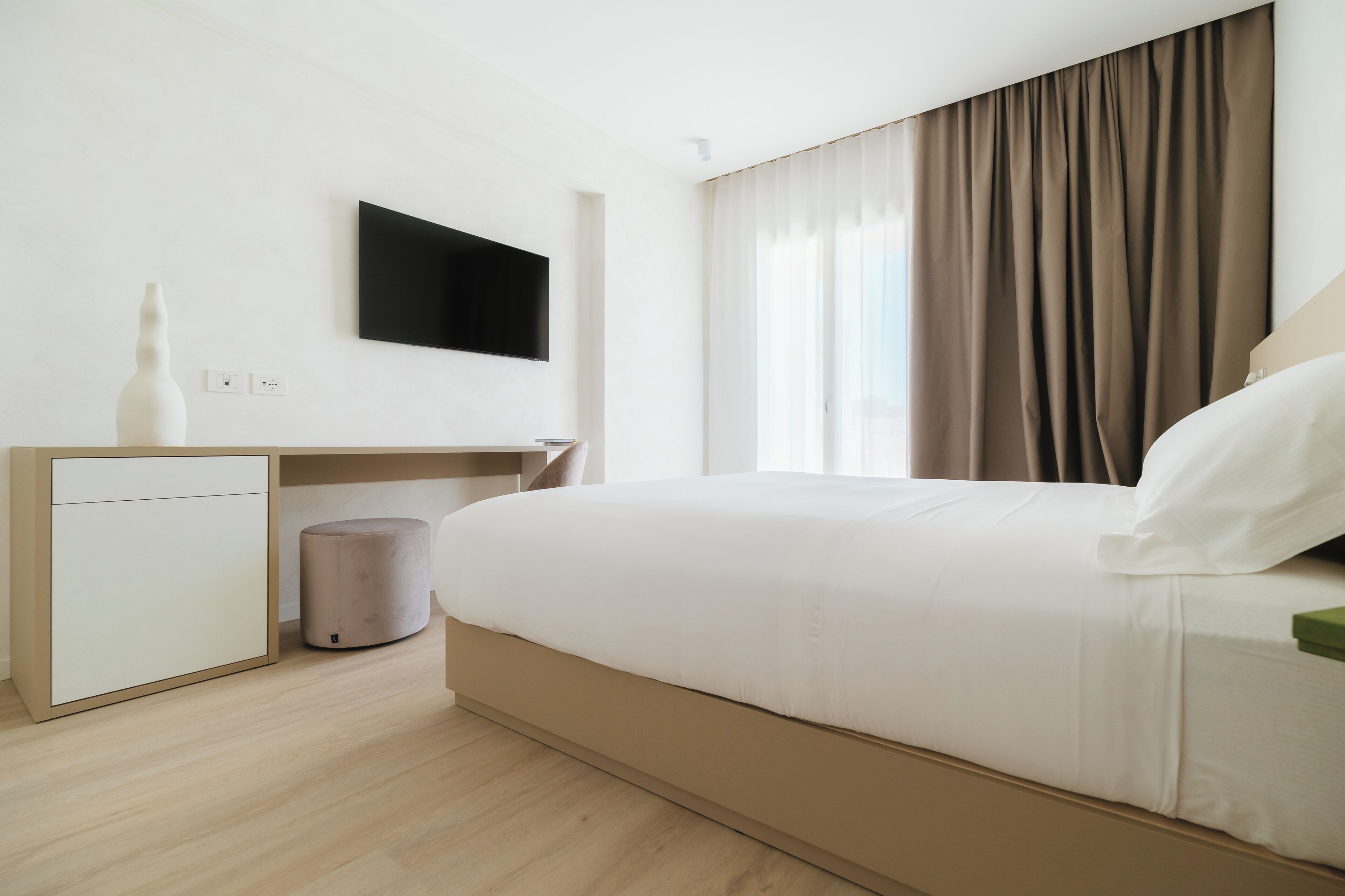 A modern hotel room with a large bed, a flat-screen TV mounted on the wall, a small desk with a vase, and a waste bin.