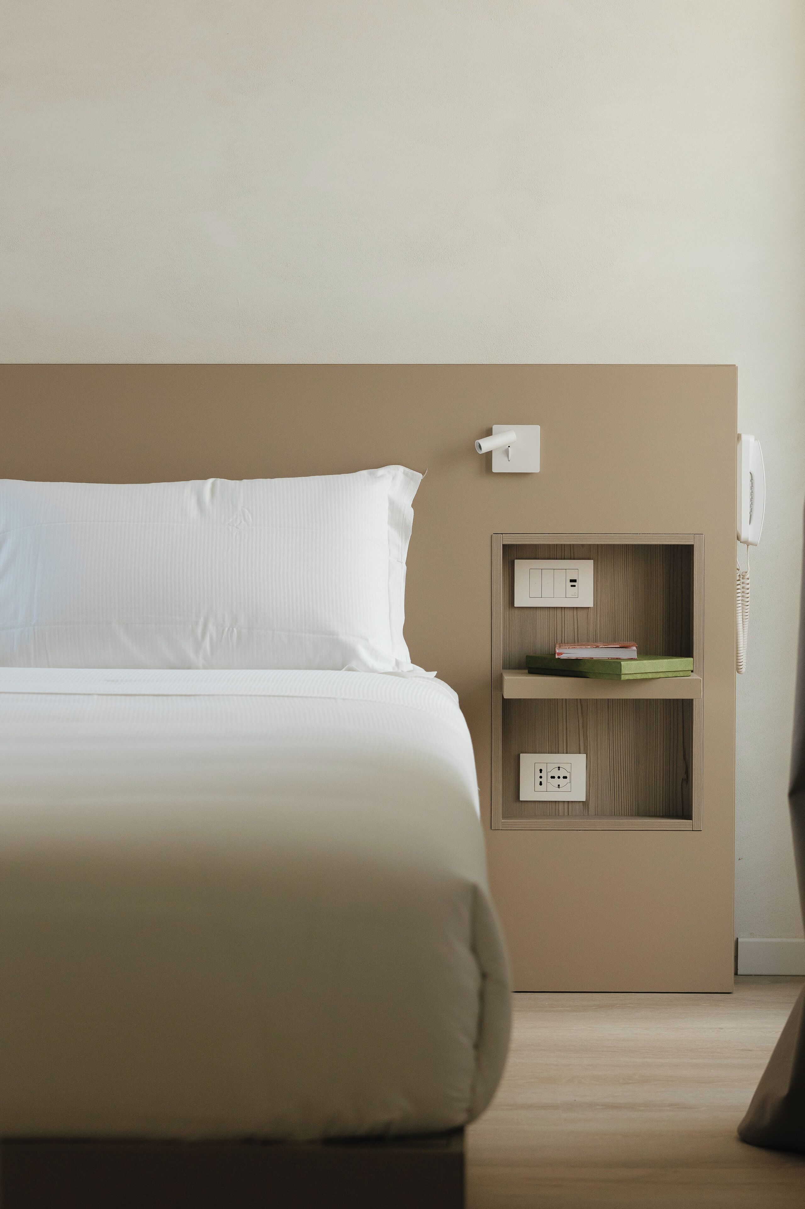 A hotel room with a bed, a bedside table with a phone, and a shelf with books.
