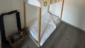 A small, cozy room with a wooden bunk bed featuring a white canopy with star decorations. A small basket with books is placed beside the bed, and a wall-mounted light fixture is visible. - Charming air-conditioned apartment (Sérignan)