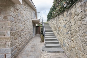 A narrow stone pathway leading to a staircase with a metal railing, flanked by stone walls and a small plant.