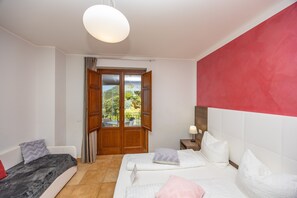 Ein Schlafzimmer mit Bett, Sofa, Nachttisch, Lampe und Blick ins Freie durch eine Tür.