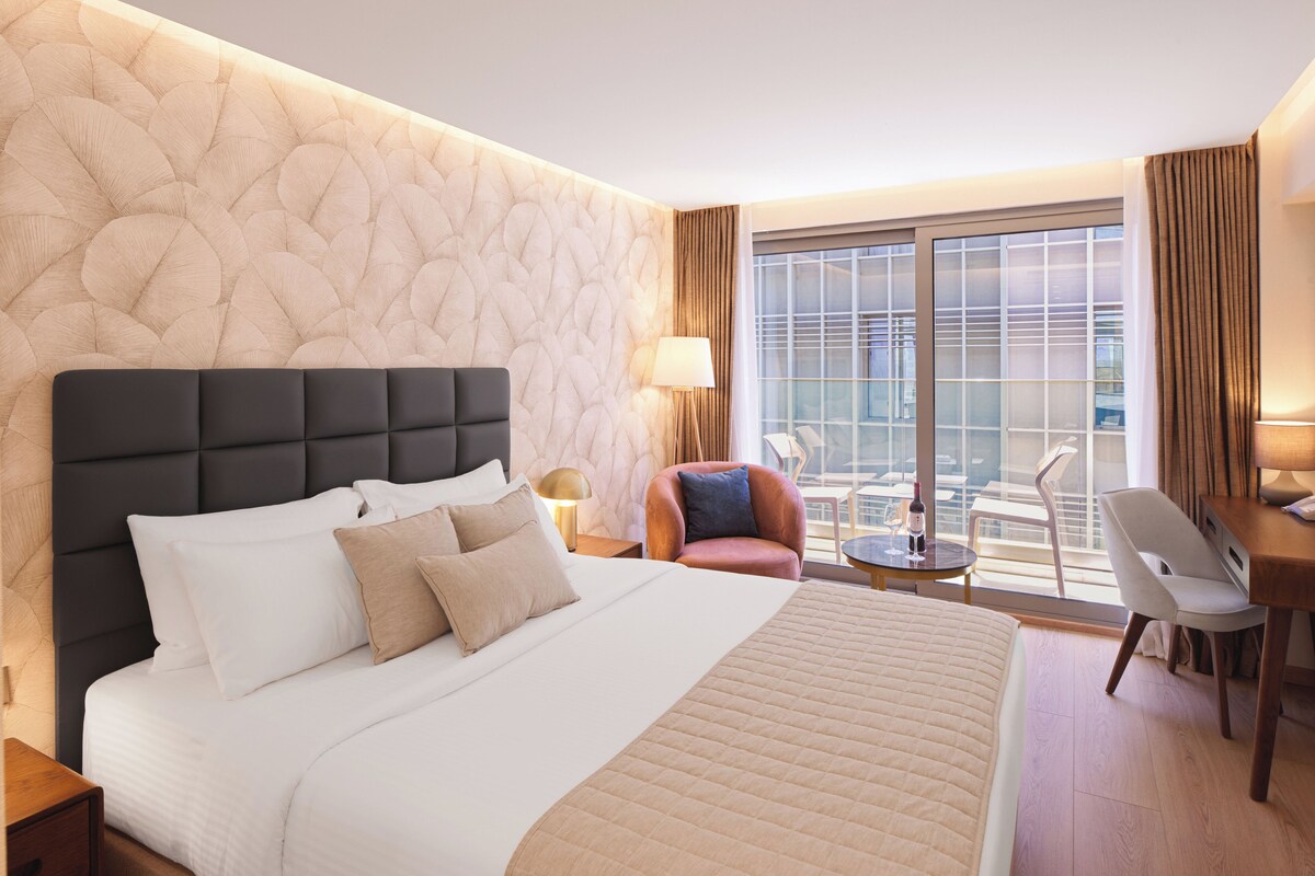 Ein Hotelzimmer mit einem großen Bett, einer Sitzecke mit Stuhl und Tisch sowie einem Ausblick durch ein großes Fenster.
