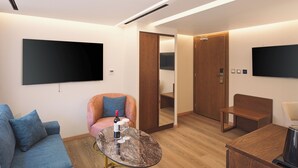 Et moderne hotelværelse med et fladskærms-tv, en sofa, et rundt sofabord med vinglas, en trædør og en træbænk.
