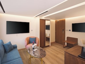 Ein modernes Hotelzimmer mit Flachbildfernseher, einer Couch, einem runden Couchtisch mit Weingläsern, einer Holztür und einer Holzbank.