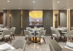 Un área de comedor moderna con mesas y sillas redondas, un aplique decorativo central y vistas del espacio de comedor a través de un panel de vidrio.