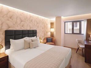 Habitación de hotel con una cama grande, cabecero, escritorio con silla, lámpara y ventana con cortinas.