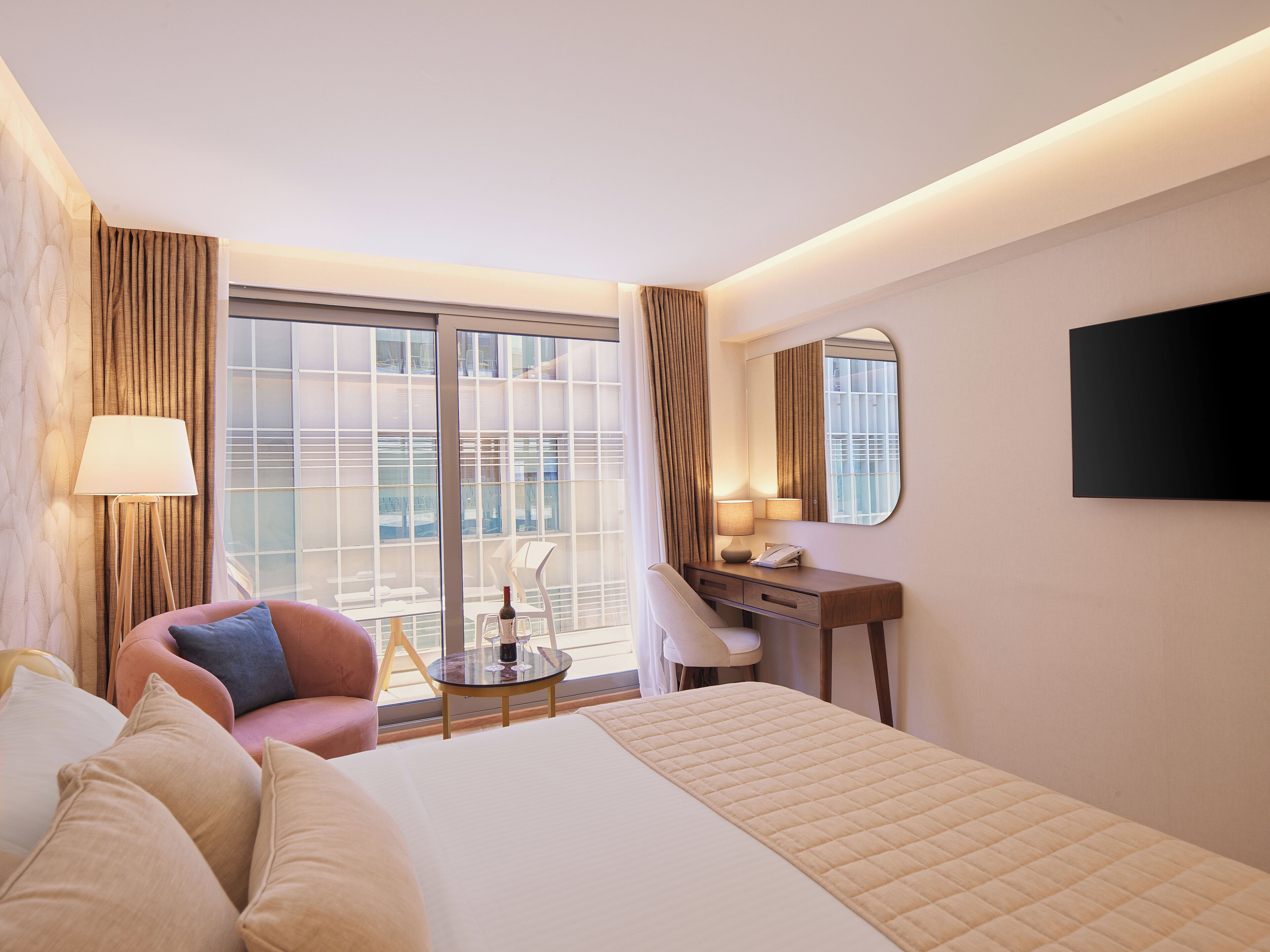 A hotel room with a bed, a desk, a chair, a lamp, a TV, and a view of the outside.