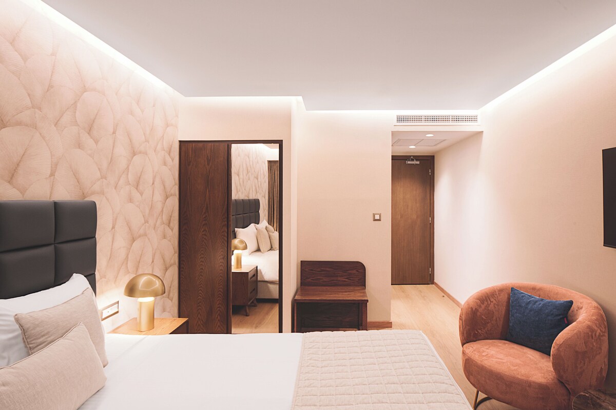 Ein modernes Schlafzimmer mit einem großen Bett, einem Nachttisch, einem Sessel, einem Holzschrank und einem durchgehenden Türspalt sichtbaren Badezimmer.