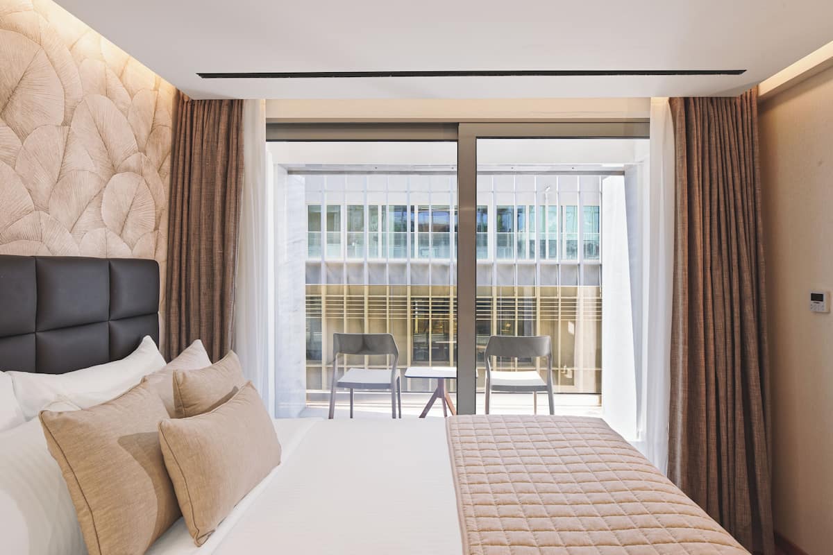 A modern hotel room with a large bed, two chairs, a balcony with a view of a building, and a control panel on the wall.