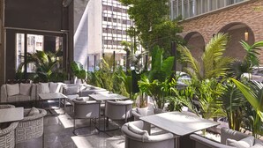 A modern outdoor seating area with multiple tables and chairs, surrounded by lush greenery and a view of a cityscape. - Leonardo Hotel Athens City Center (Athens)
