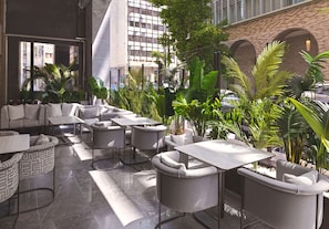 A modern outdoor seating area with multiple tables and chairs, surrounded by lush greenery and a view of a cityscape.