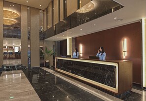 Ein Hotel-Empfangsbereich mit einer Marmor-Theke, zwei Rezeptionisten und einer Bar im Hintergrund.