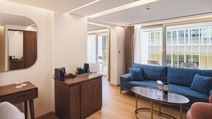 Et moderne hotelværelse med et træskrivebord, en sofa, et glasbord og et stort vindue med udsigt over byen.