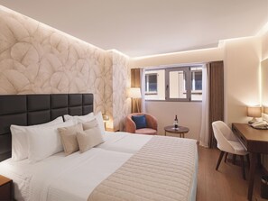 A hotel room with a large bed, a desk, a chair, a small table with a wine bottle, and a window with curtains.