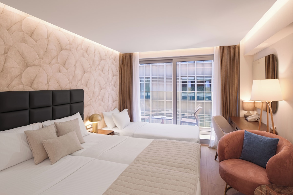 Ein Hotelzimmer mit einem großen Bett, einem kleineren Bett, einer Couch, einem Sessel, einem Schreibtisch, einer Lampe und einem fenster mit Vorhängen.
