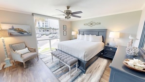 A bedroom with a large bed, a chair, a bench, a nightstand, a lamp, and a decorative item on the nightstand. - Shoreline Towers 3025 2 Bedroom Condo (Destin)