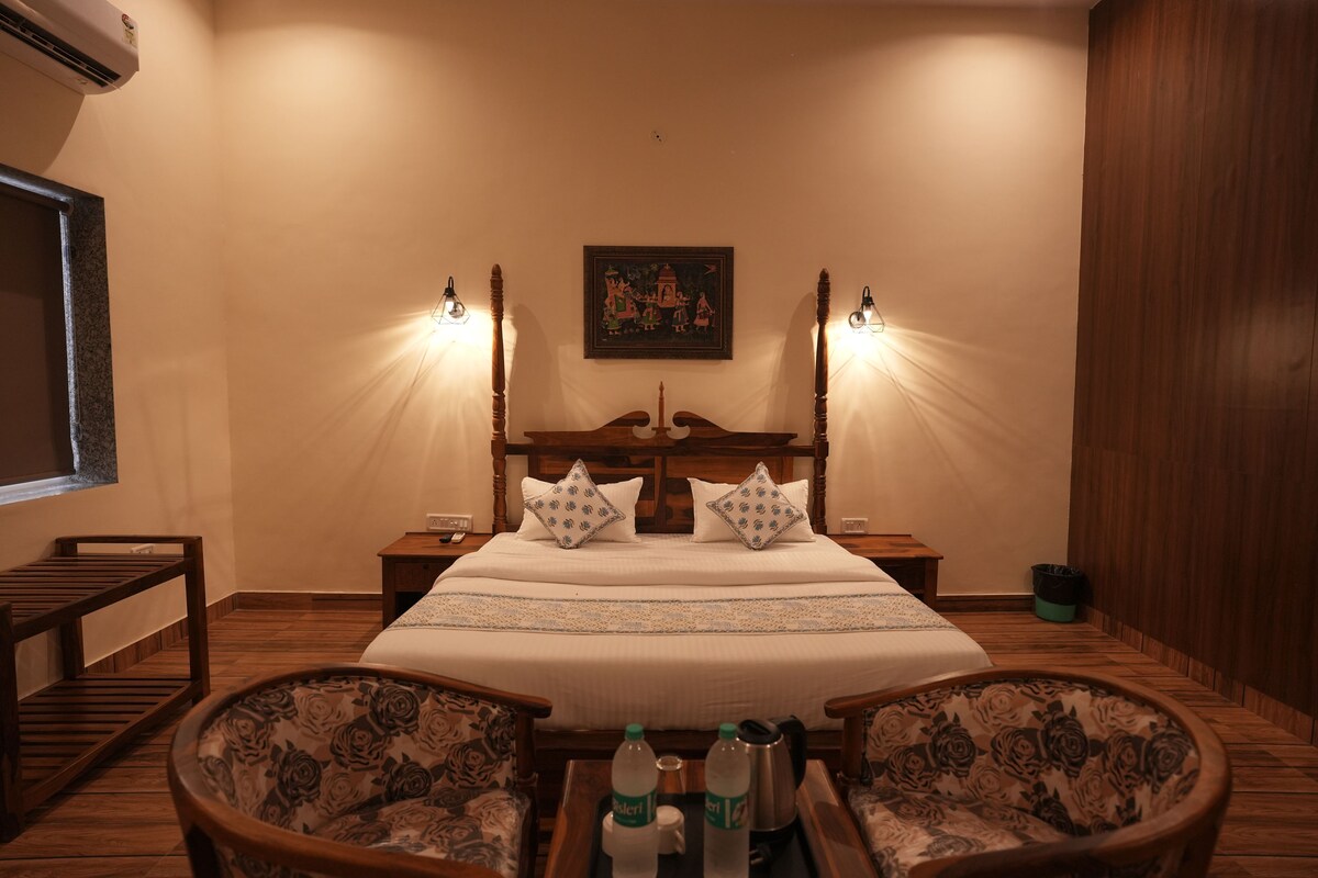 A hotel room with a large bed, two bedside tables, a wooden bench, two armchairs, and a wall-mounted air conditioner.