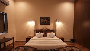 A hotel room with a large bed, two bedside tables, a wooden bench, two armchairs, and a wall-mounted air conditioner.