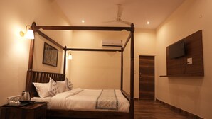 A four-poster bed with a wooden frame, a bedside table with a lamp, a flat-screen TV mounted on the wall, and an air conditioning unit above the bed.