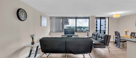 Ein modernes Wohnzimmer mit einer Couch, einem Fernseher und Blick auf die Stadt durch große Fenster.
