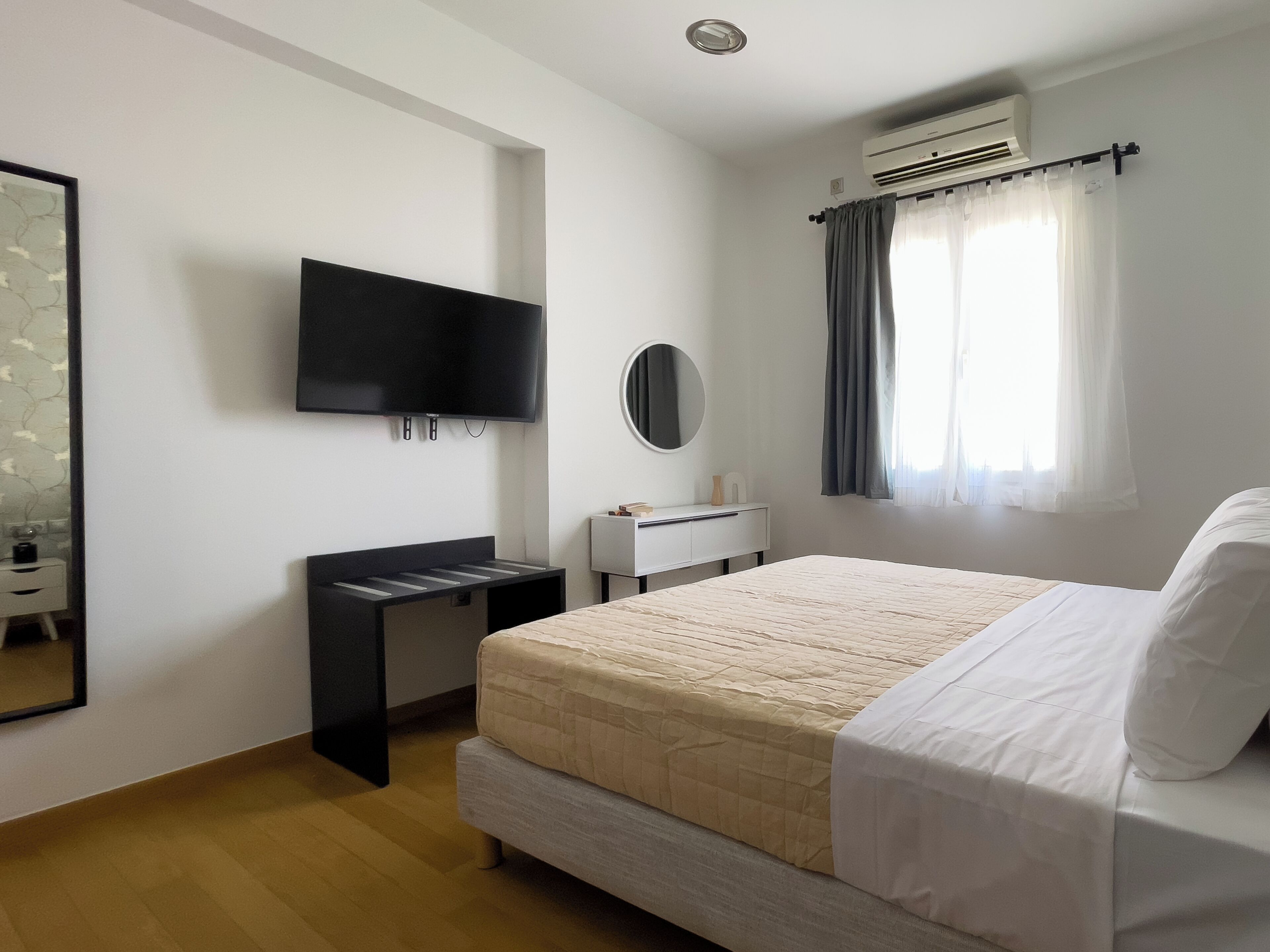 Ein Schlafzimmer mit einem Bett, einem an der Wand befestigten Fernseher, einem Schreibtisch, einem Spiegel und einer Klimaanlage.