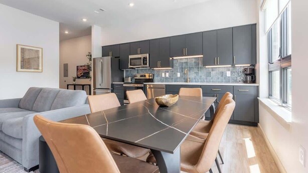 A modern kitchen with dark cabinetry, a central island, and a dining area with a table and chairs. - Music Row Magic - Pool + Gym + Downtown (Nashville)