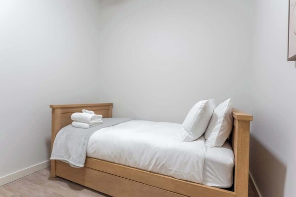 Una cama individual con estructura de madera, ropa de cama blanca y una toalla doblada al lado.
