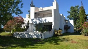 Un edificio blanco de dos pisos con un balcón en el nivel superior, rodeado de follaje y árboles.