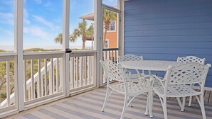 A covered outdoor area with a white table and chairs, a view of the beach, and a building with a blue exterior.