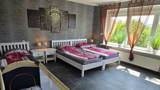 A bedroom with two beds, a crib, a large window with curtains, and a wall-mounted artwork.