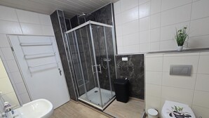 A modern bathroom with a glass-enclosed shower, a freestanding bathtub, a toilet with a decorative cover, and a small potted plant on the wall.