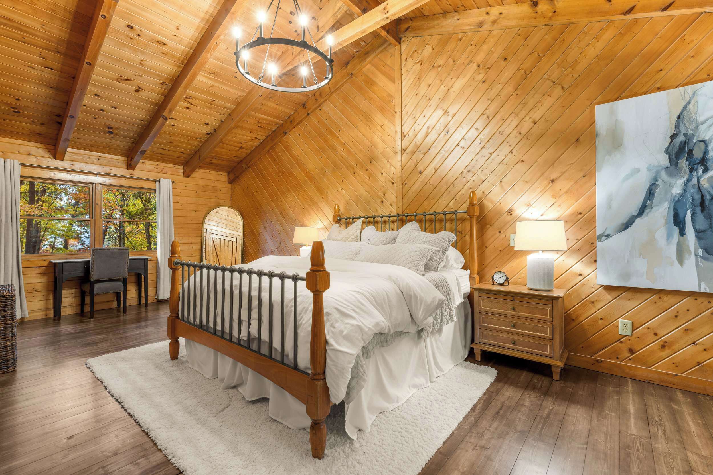 A cozy bedroom with a wooden ceiling, a bed with white linens, a bedside table with a lamp, and a large abstract painting on the wall.