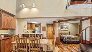 Une cuisine spacieuse avec des meubles de rangement en bois et un coin repas comprenant une table et des chaises en bois. La pièce adjacente contient un lit recouvert d’une courtepointe matelassée.