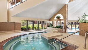 Un bain à remous bordé de briques, avec une échelle en métal, et une vue sur un jardin avec une piscine et de la verdure.