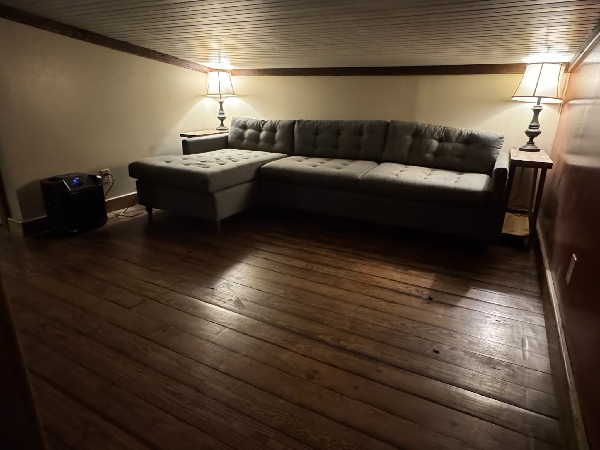 Una habitación con suelo de madera, un sofá modular gris, dos lámparas y un pequeño altavoz negro.
