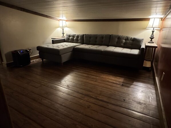 A room with a wooden floor, a grey sectional sofa, two lamps, and a small black speaker.