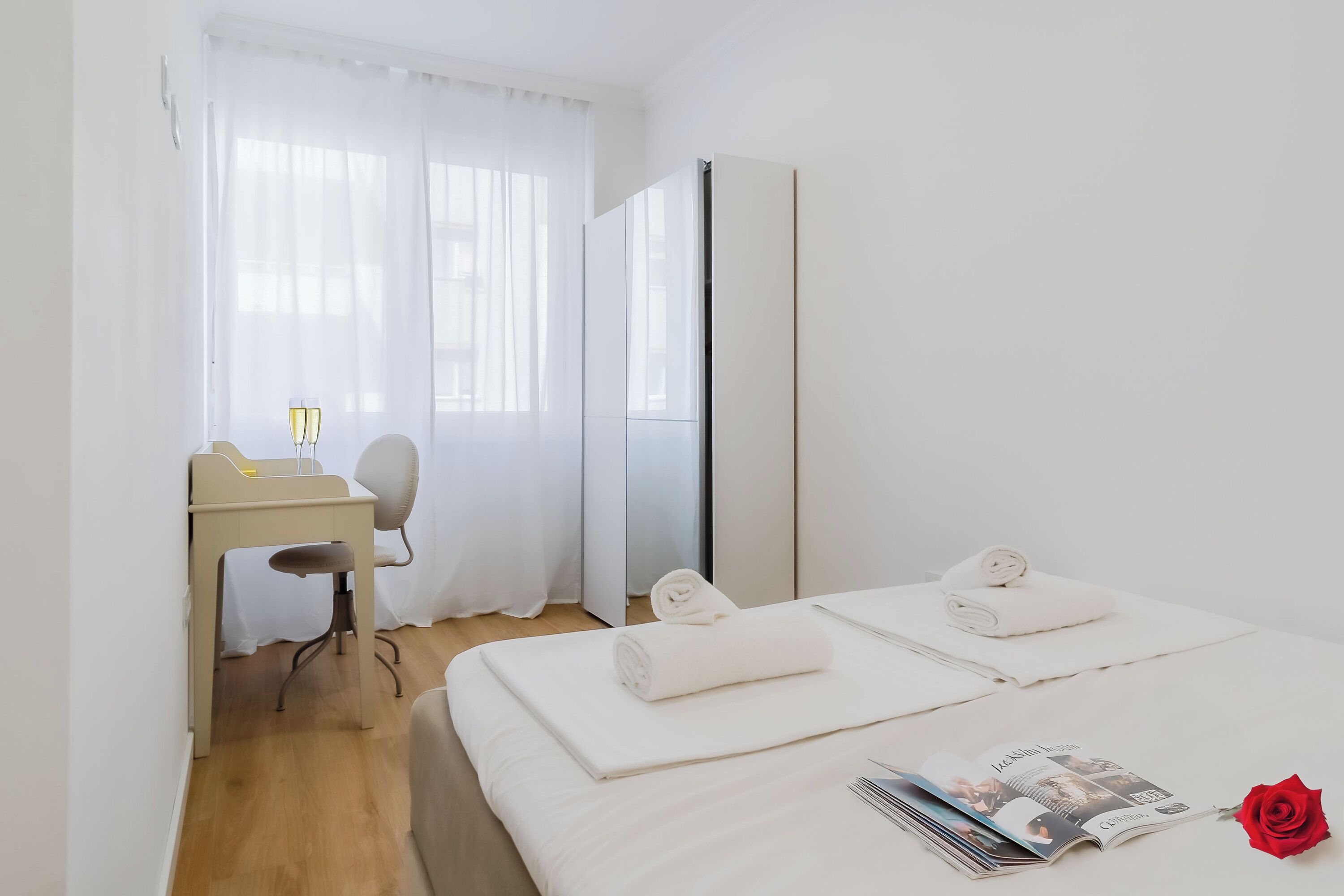Een hotelkamer met een bed, een bureau, een stoel, een spiegel en een rode roos.