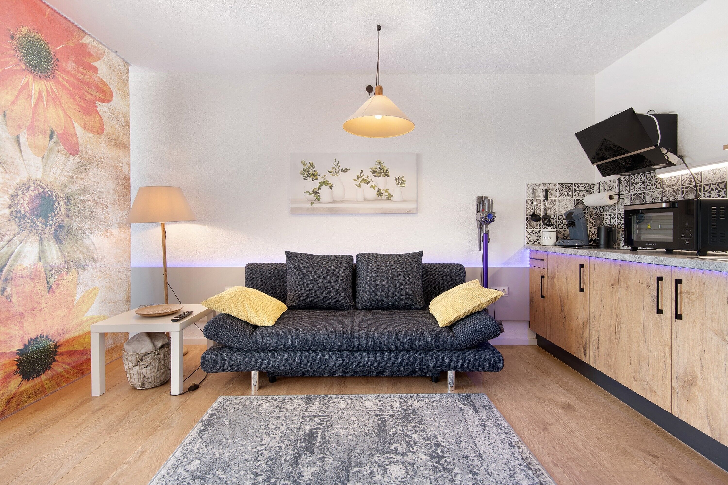 Uma sala de estar moderna com um sofá, um abajur, um quadro e uma área de cozinha com armários e eletrodomésticos.