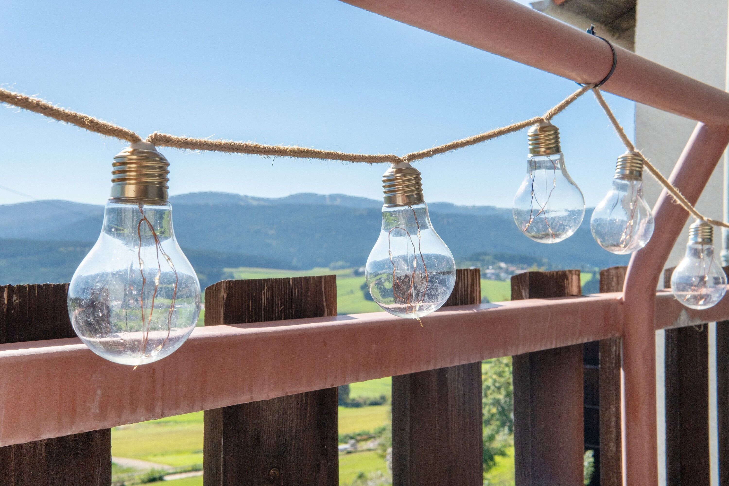 A string of light bulbs hanging on a wooden railing with a view of green fields and mountains in the background.