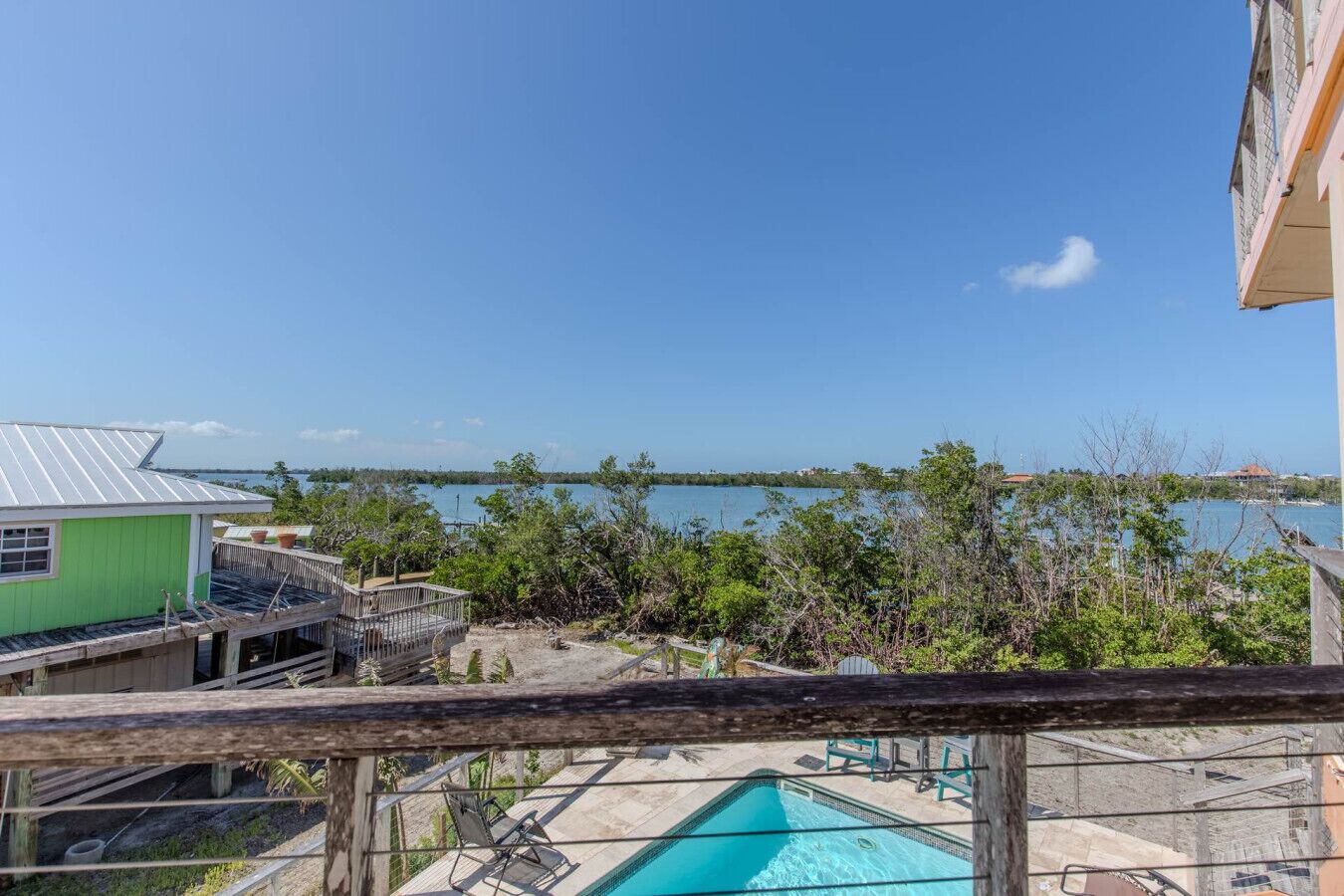 A view of a swimming pool with a wooden deck and lounge chairs, surrounded by greenery and overlooking a body of water.